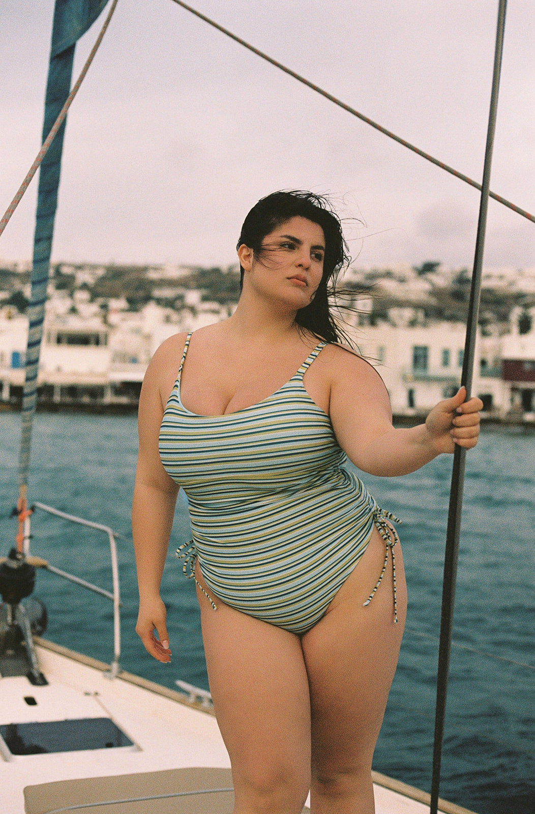 Sorbet ruched one-piece swimsuit with blue and yellow stripes, side ties, and spaghetti straps, worn on a sailboat in a lifestyle shot.