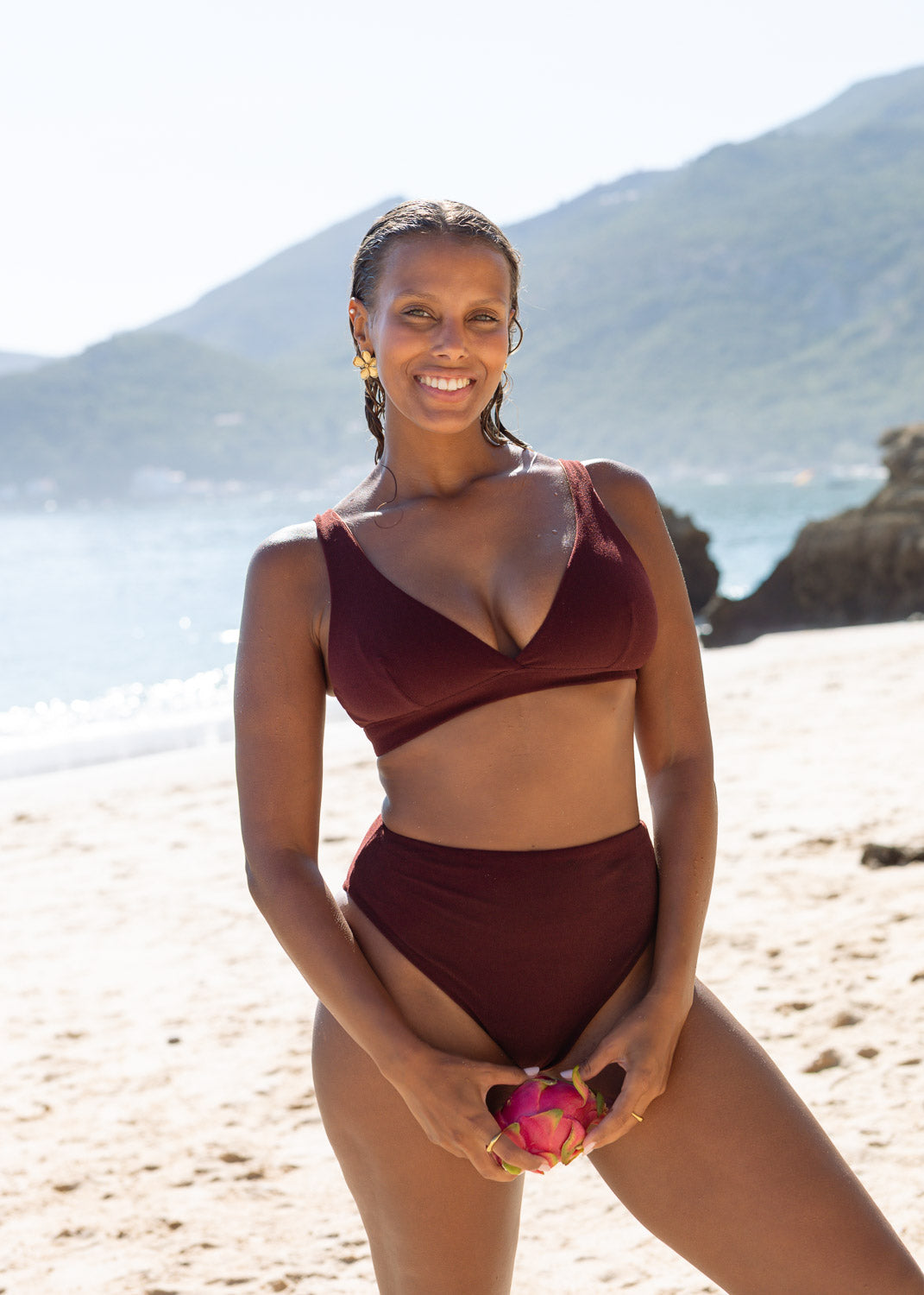 Maroon high-leg bikini bottoms by Lilja the Label, worn with a matching top, and gold earrings, on a beach.