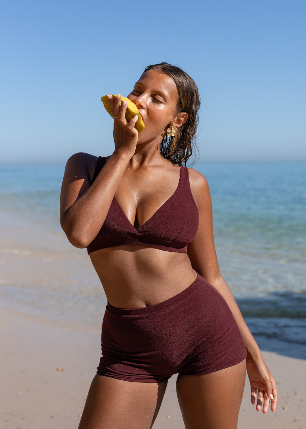 Burgundy plunge top by Lilja the Label, worn with matching bottoms, with a woman eating a yellow fruit.