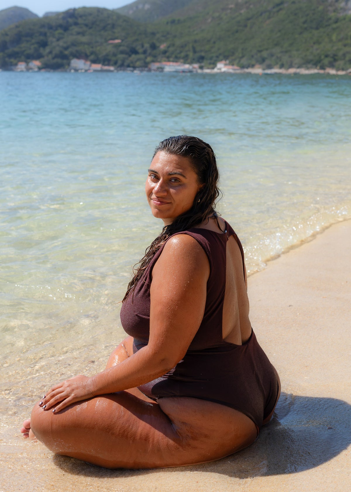 Brown boatneck onepiece swimsuit with open back, worn by a woman seated on a sandy beach.