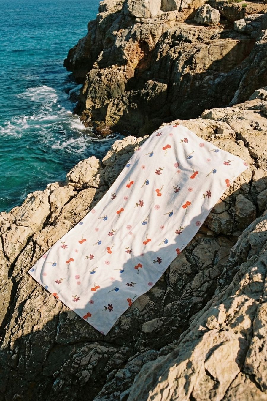 Floral bath towel by Lilja the Label on tan rocks near the ocean.
