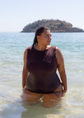 Maroon boatneck one-piece swimsuit by a model sitting in shallow ocean water, showcasing its smooth texture and fitted silhouette with a side view.