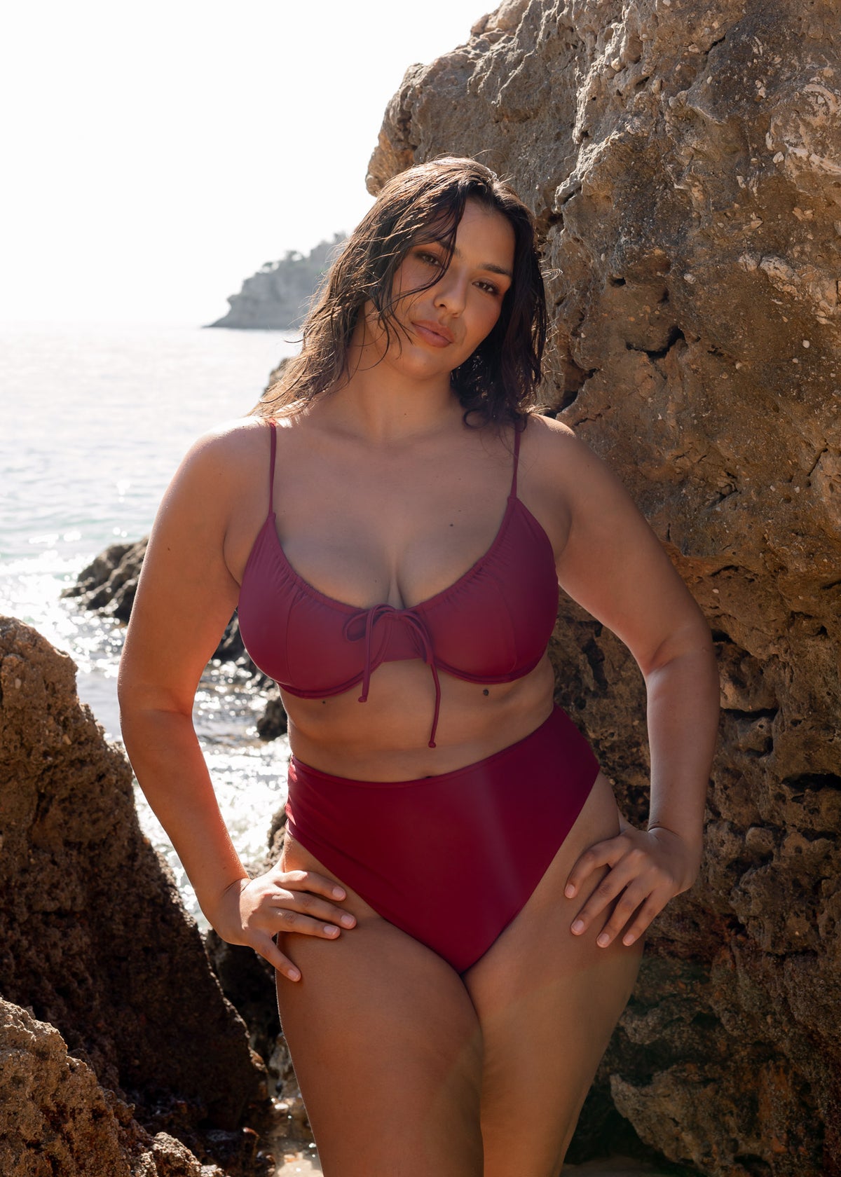 Bordeaux high-waisted midi bikini bottoms modeled against coastal rocks, paired with a matching strappy top.