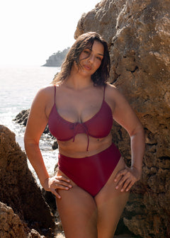 Bordeaux high-waisted midi bikini bottoms modeled against coastal rocks, paired with a matching strappy top.